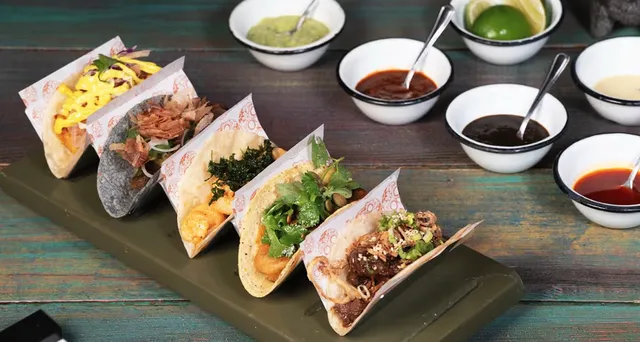 Five tacos lined up in paper sleeves on a wooden board, with small bowls of salsa and lime wedges behind them.