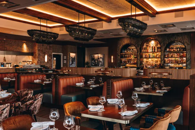 Dining room with tan leather booths, set tables, woven pendant lights and a backlit arched bar lined with bottles.
