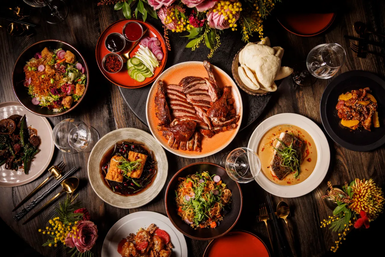 Overhead spread of Chinese dishes on a dark wood table, with sliced duck at the center, steamed bao, noodles, dumplings and flowers.