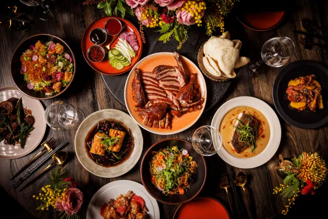 Overhead spread of Chinese dishes on a dark wood table, with sliced duck at the center, steamed bao, noodles, dumplings and flowers.