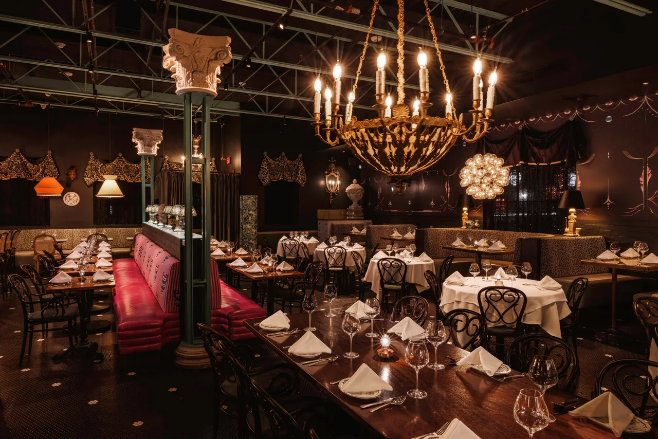Dining room under a candle chandelier, with a green column, red leather banquettes and tables set with white napkins.